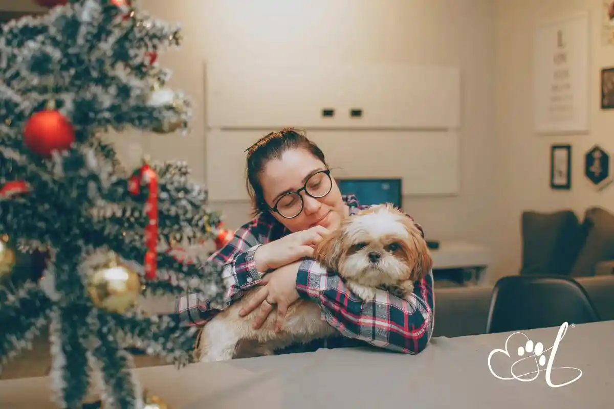 Woman in glasses gently hugs her small dog beside a decorated Christmas tree, capturing a quiet moment of love and comfort during the holiday season.